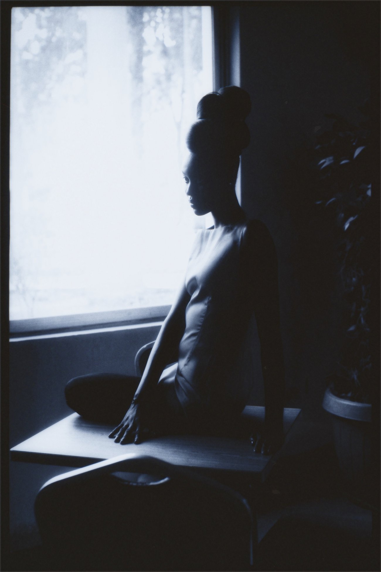 Black woman with hair in an updo sitting on a square desk in front of a bright window looking off to the side