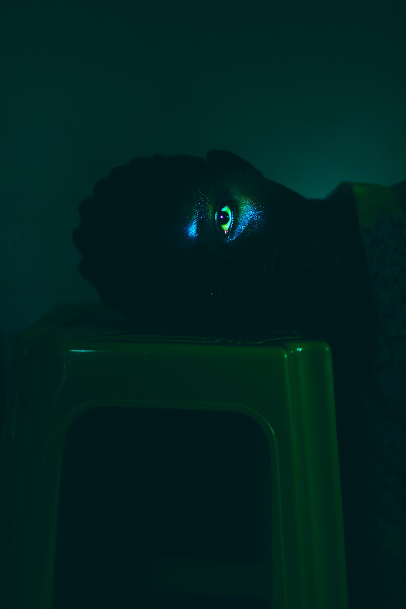 Black woman leaning head sideways on green stool with only one eye illuminated