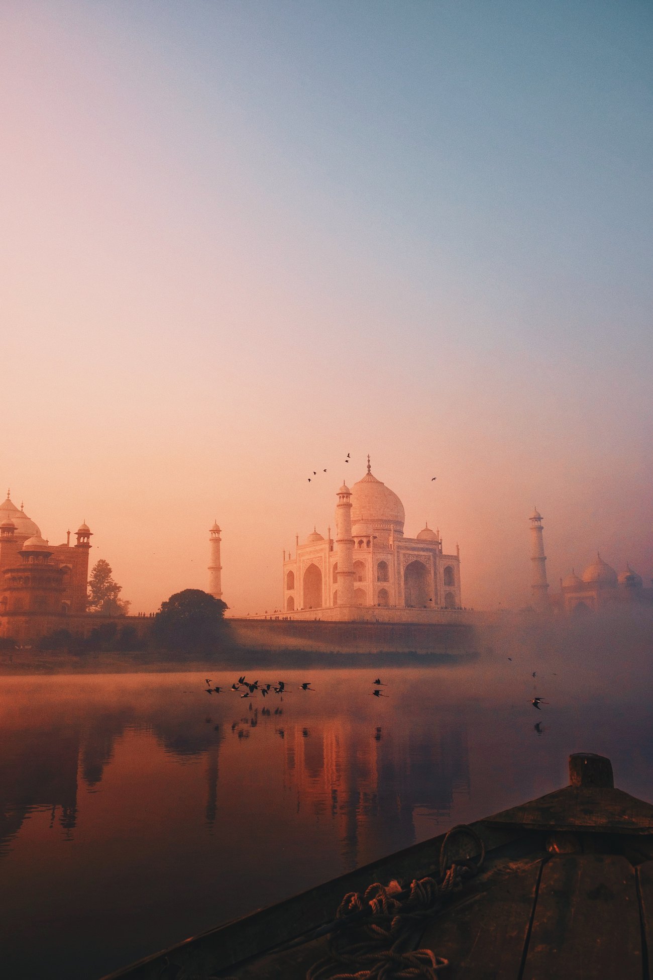 Flocks of birds flying in front of the Taj Mahal in India with peach and blue hues are seen from boat in the Yamuna river