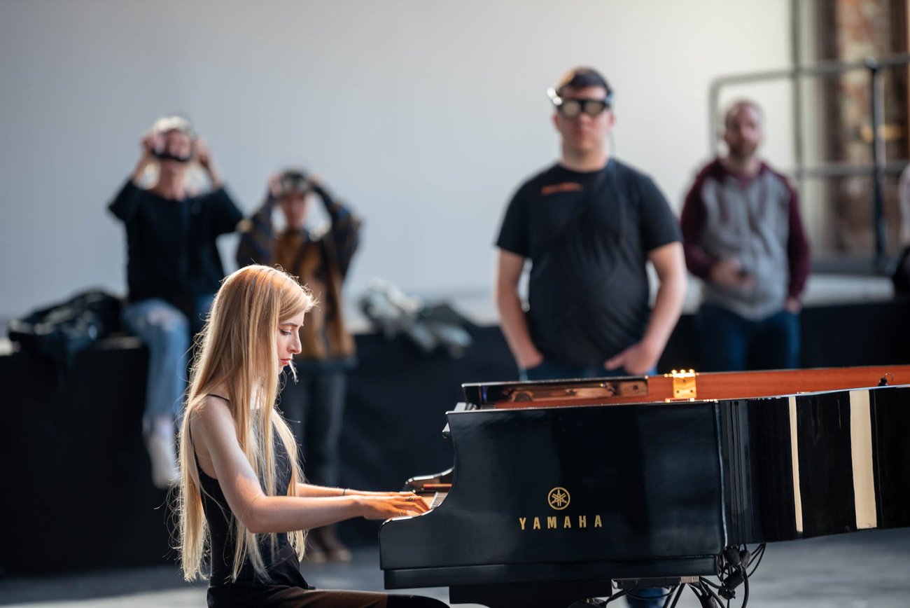 Side view of pianist Kelly Moran playing piano with people standing in background wearing viewing glasses