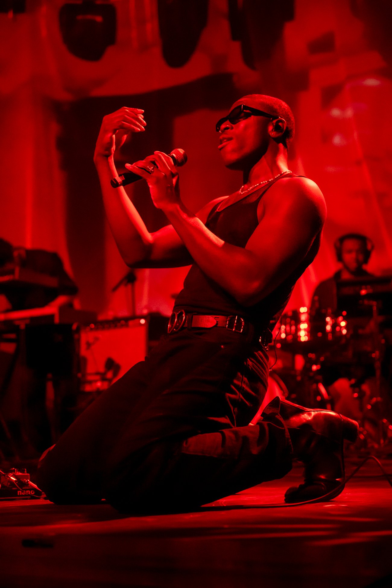 Musician Moses Sumney singing into a microphone kneeling on red lit stage while dressed in black jeans and vest