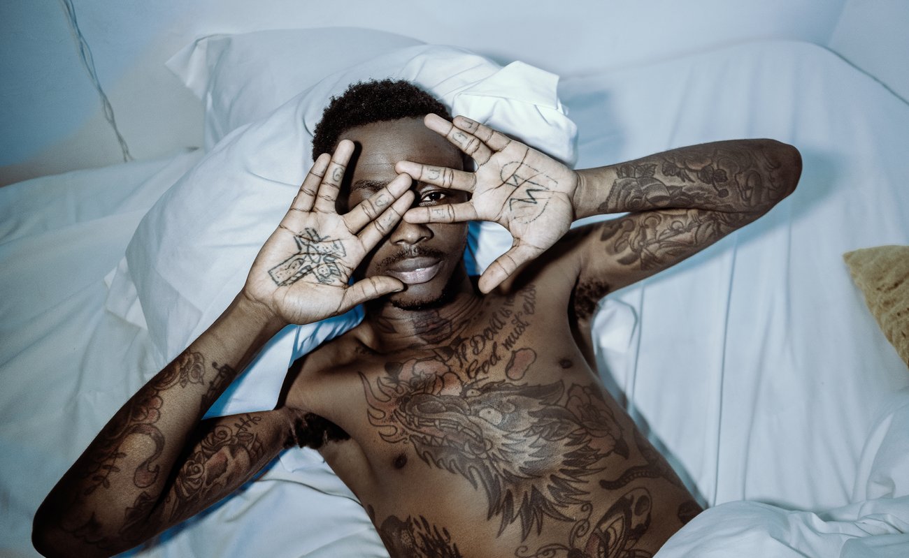 Tattooed Black man lying in white sheets with one eye looking at camera through fingers