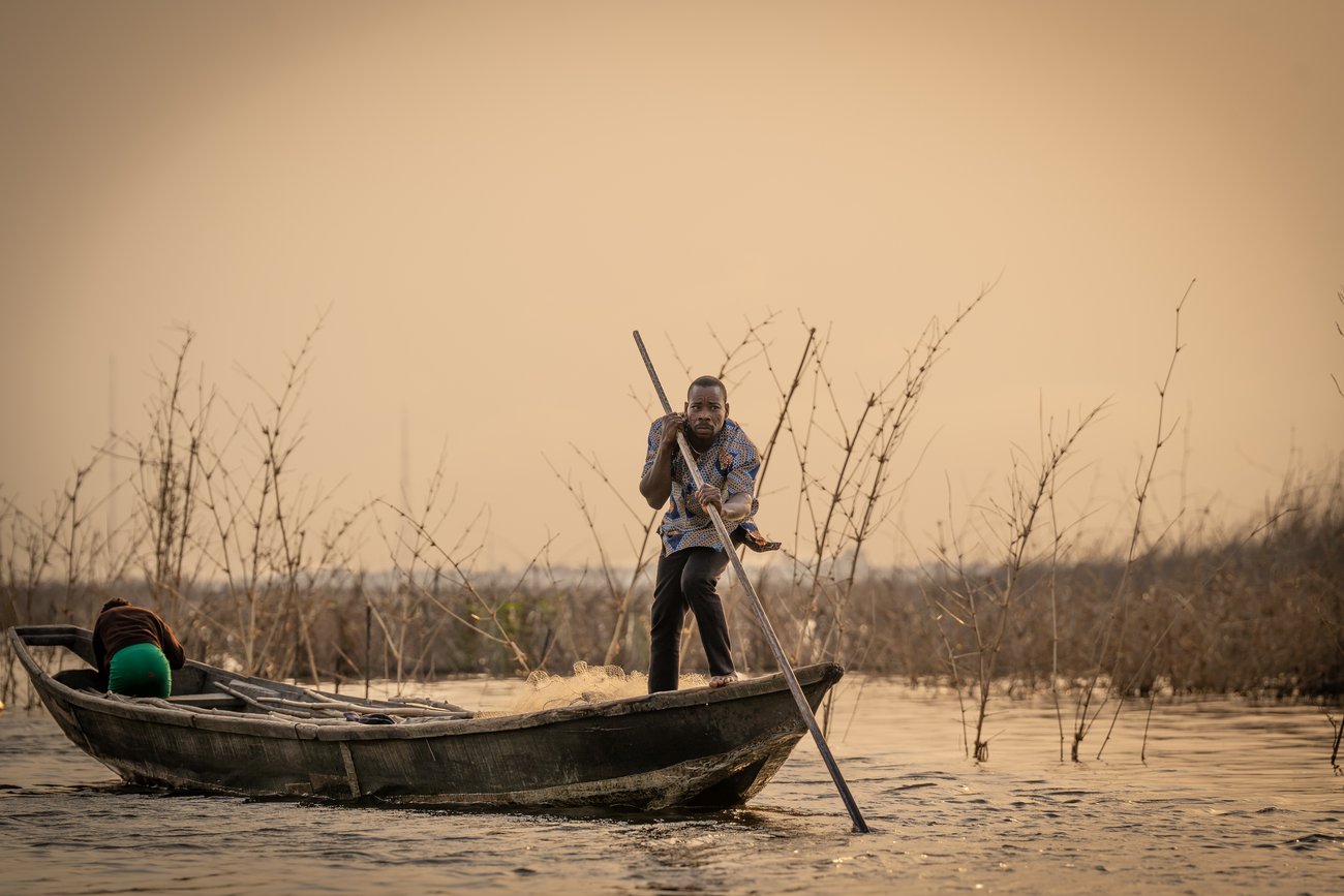 Black man standing and steering boat in shallow water surrounded by reeds