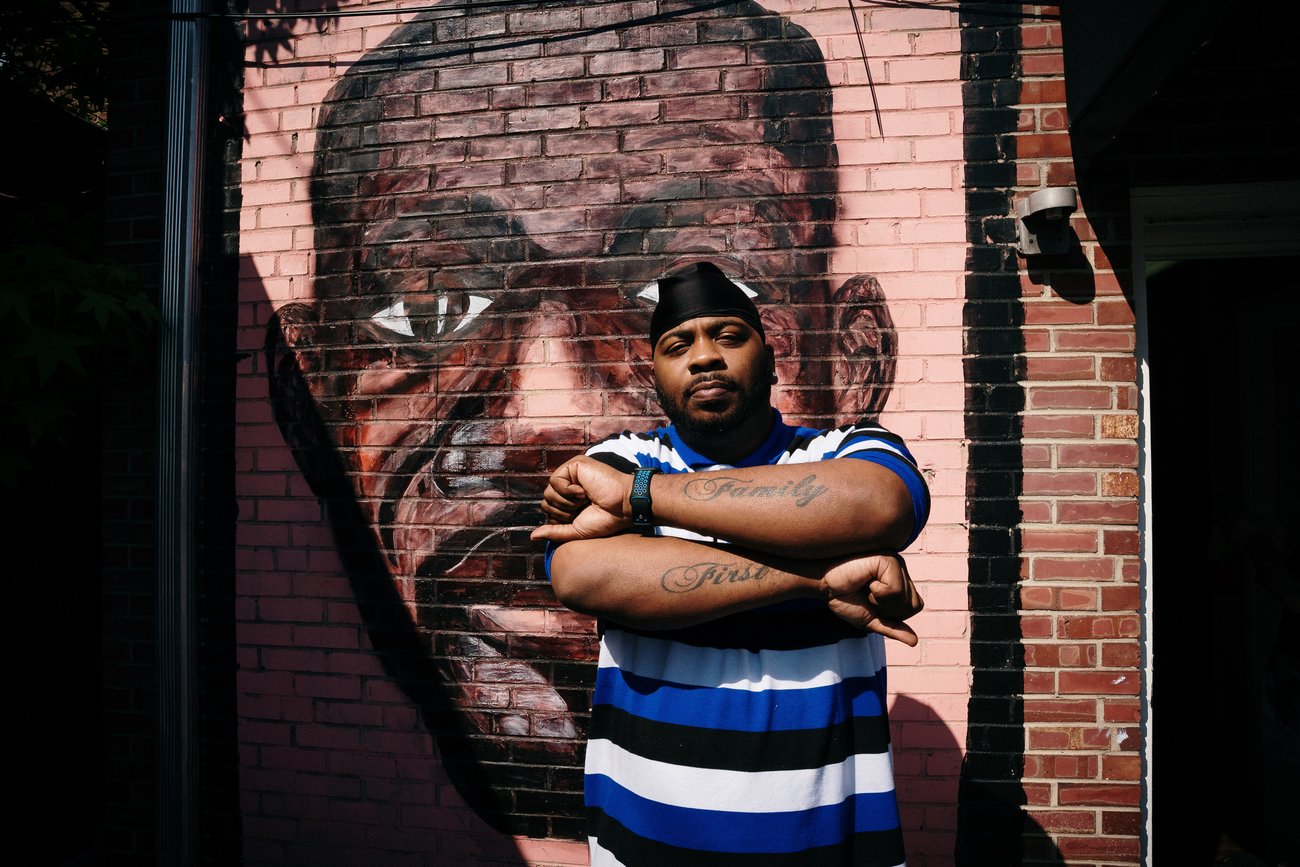 Portrait of Black man showing “family first” forearm tattoos in front of George Floyd mural on red brick wall