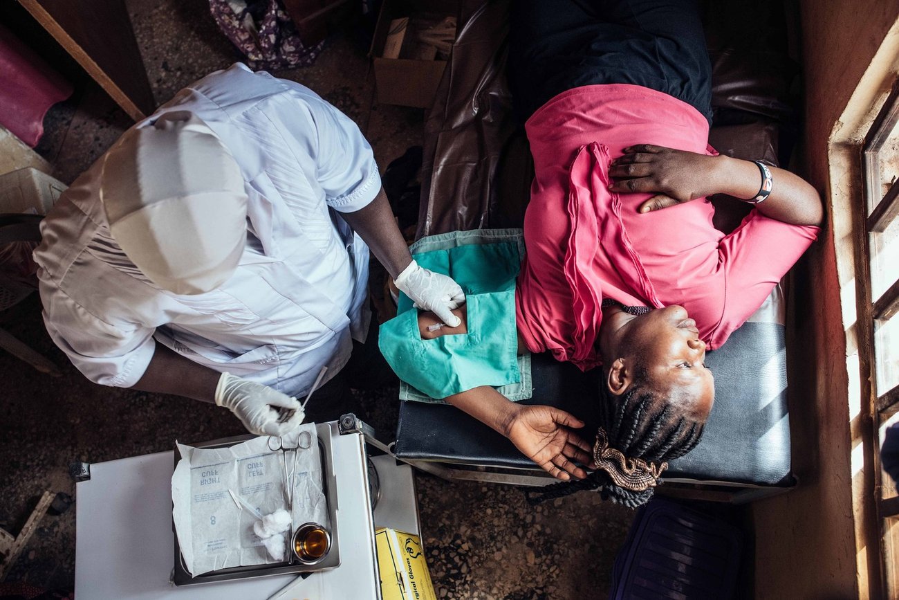 Top view of Black woman lying down looking out of window while medic works on her arm