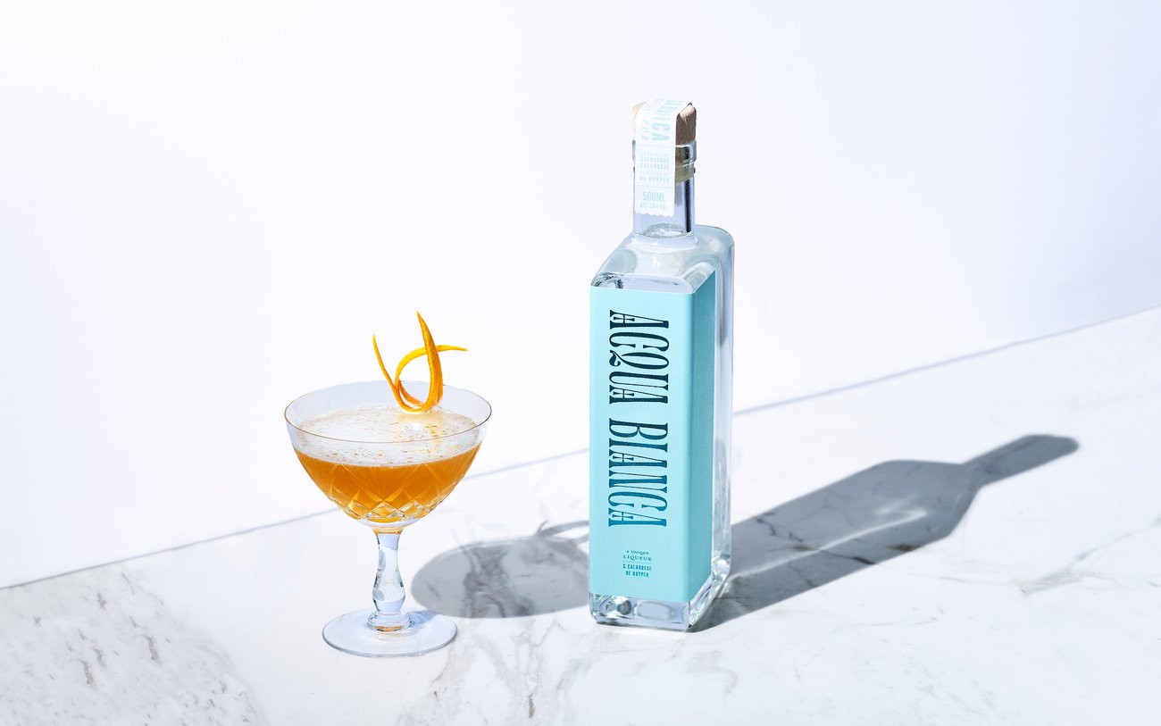 Bottle of liquor with light blue label that reads “Agua Bianca” next to an orange colored cocktail in crystal glass on marble counter with shadow