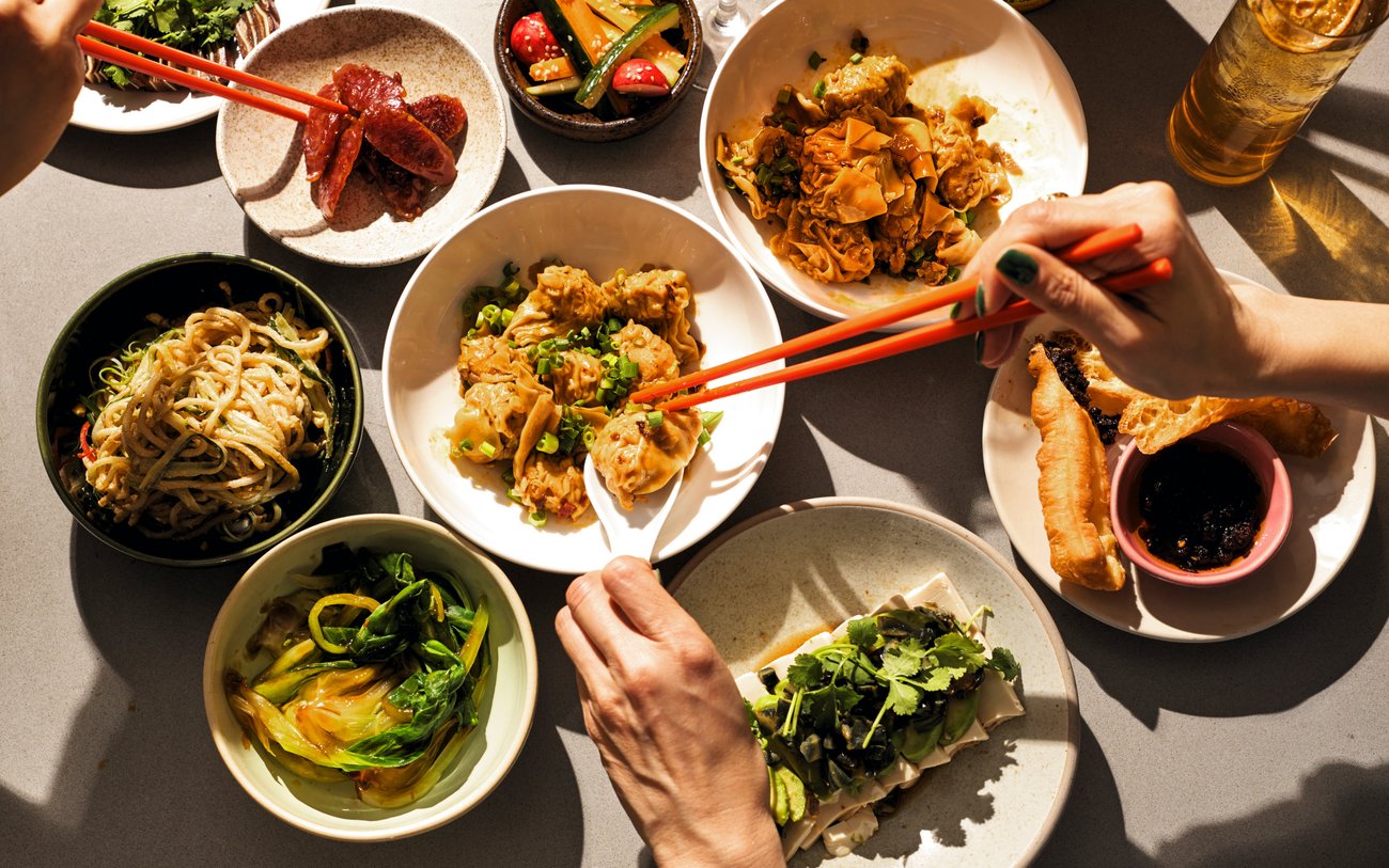 Top view of table of Asian dishes with woman’s hands reaching for dumplings with red chopsticks