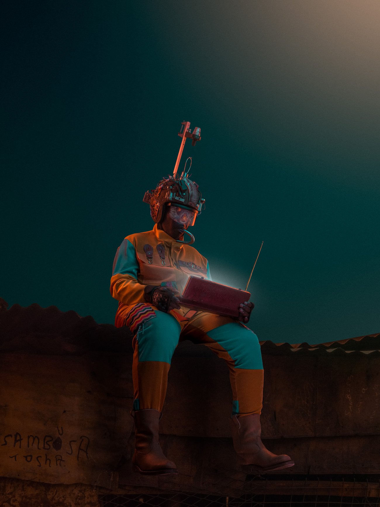 Man in metal helmet sitting on a metal roof watching a broadcast on a rectangular device under a clear night sky
