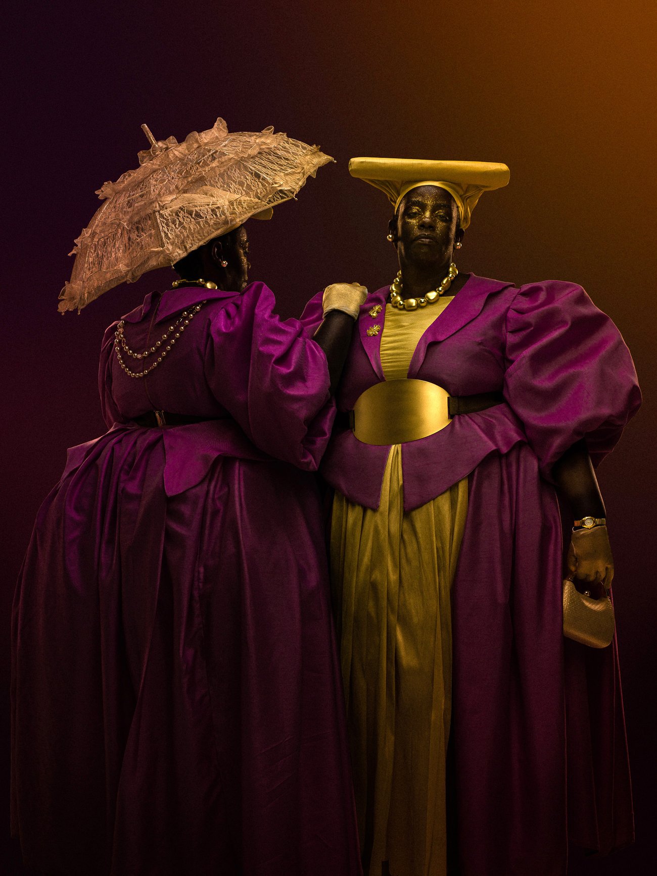 Two Black people dressed in wide oversized magenta gowns and gold accessories
