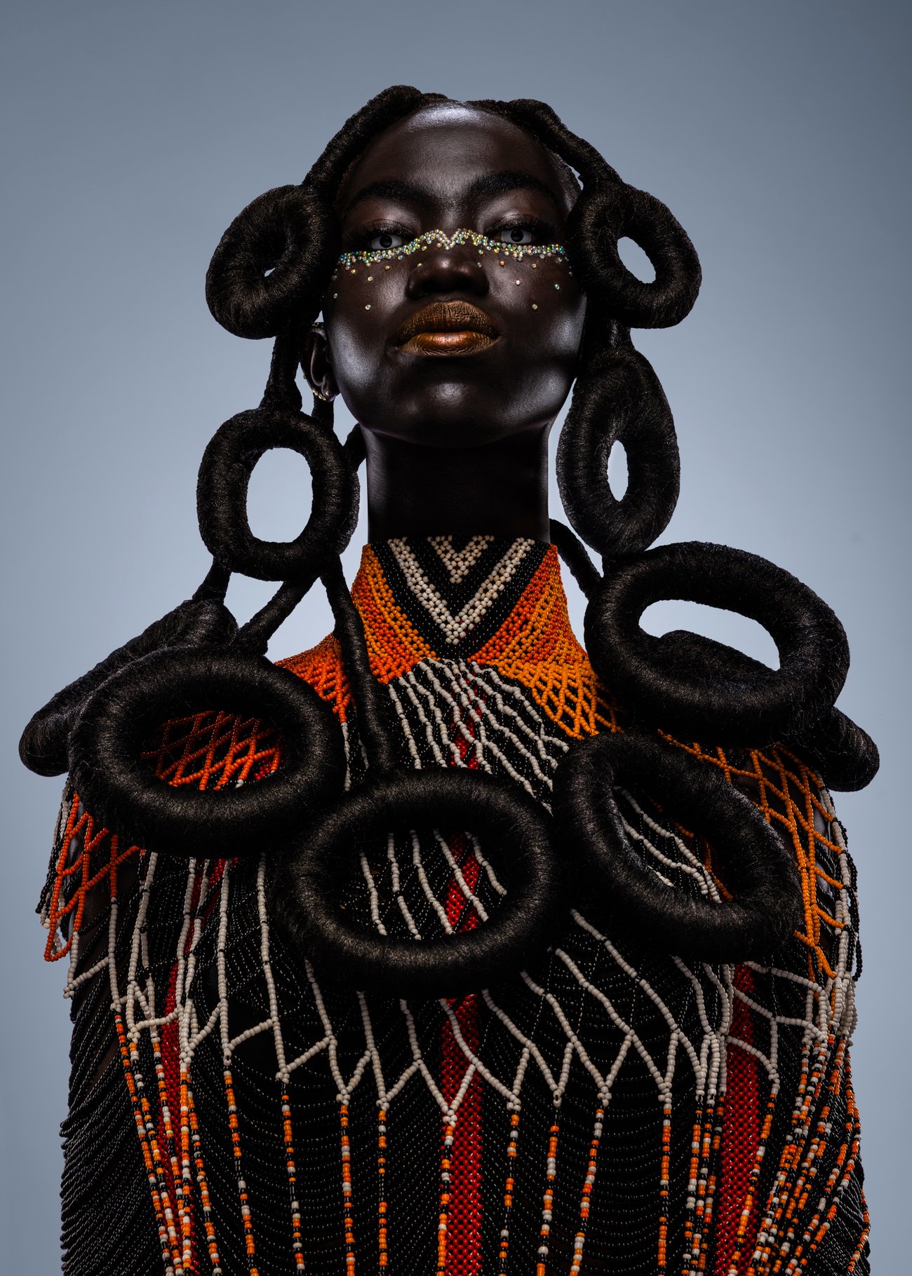 Black woman with white contact lenses and row of decorative crystals under her eyes in orange beaded top with hair designed in multiple circles