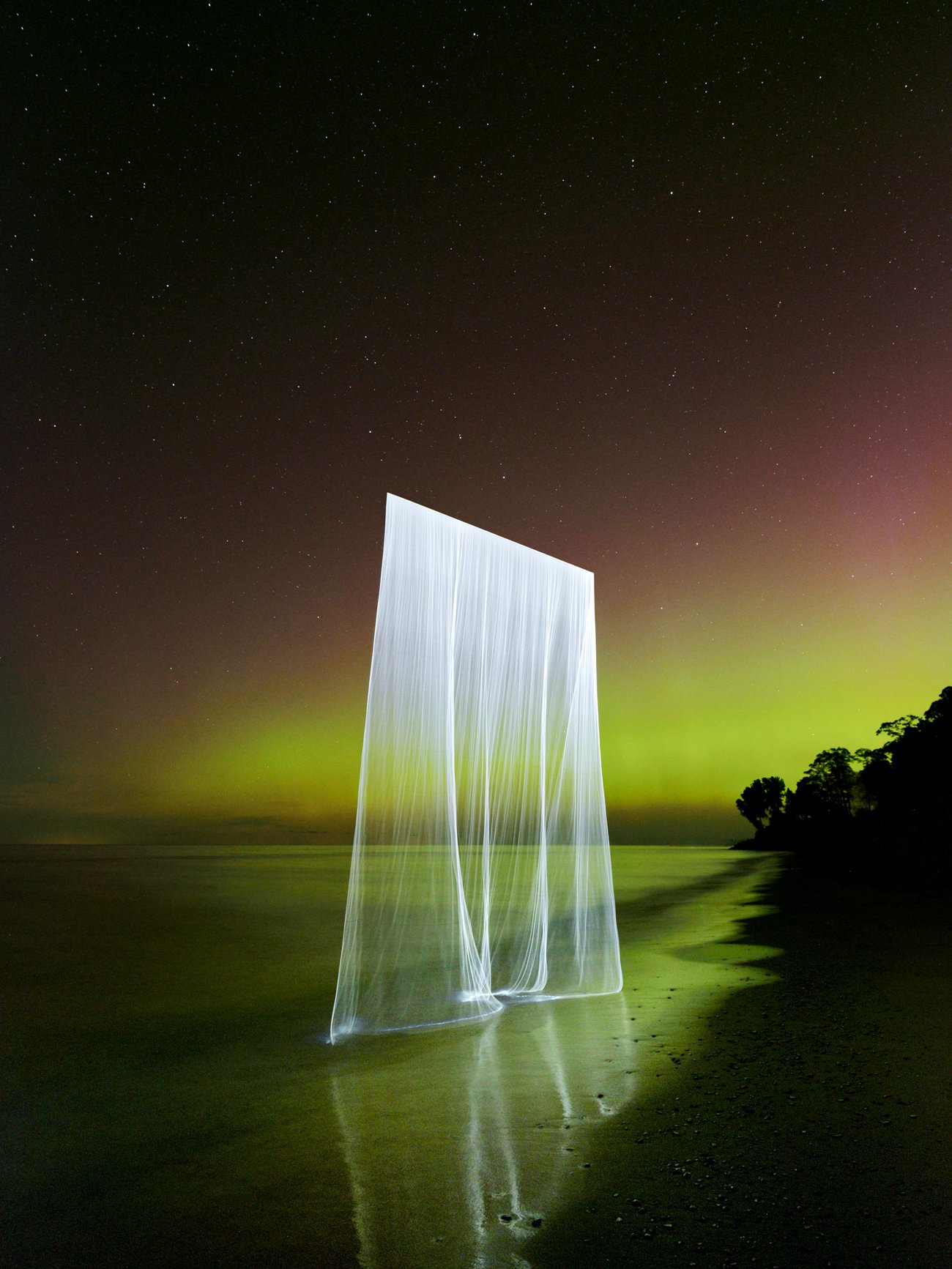 Multiple threads of thin light giving the effect of a single white translucent curtain above a dark green shoreline with the Northern Lights in the background