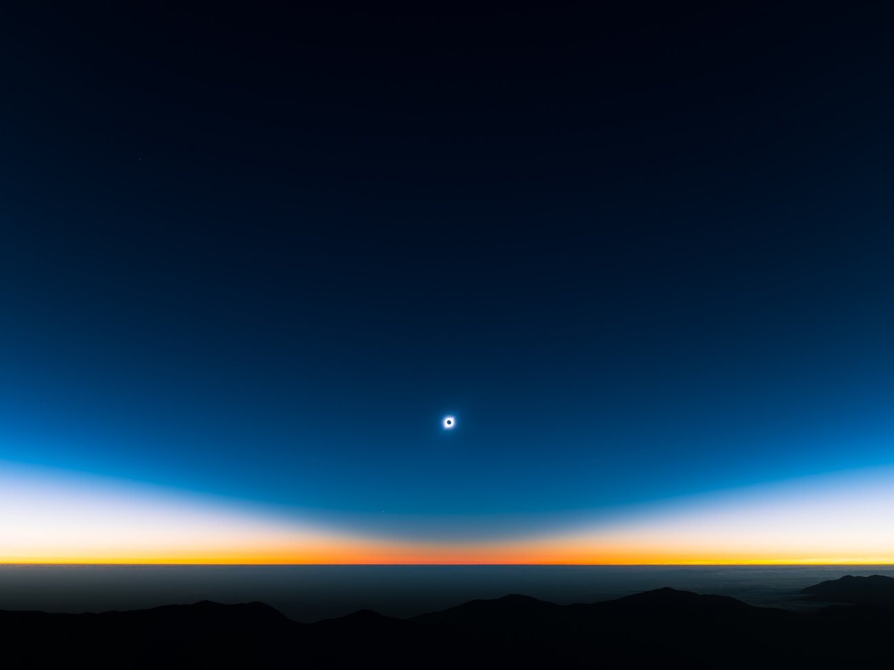 Full solar eclipse as a small circle of light in the distance of a dark gradient blue sky with yellows and oranges on the horizon