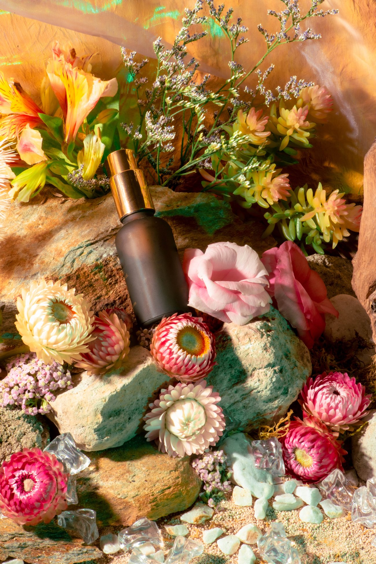 Brown glass bottle on red rocks surrounded by strawflowers, lilies, roses and succulents
