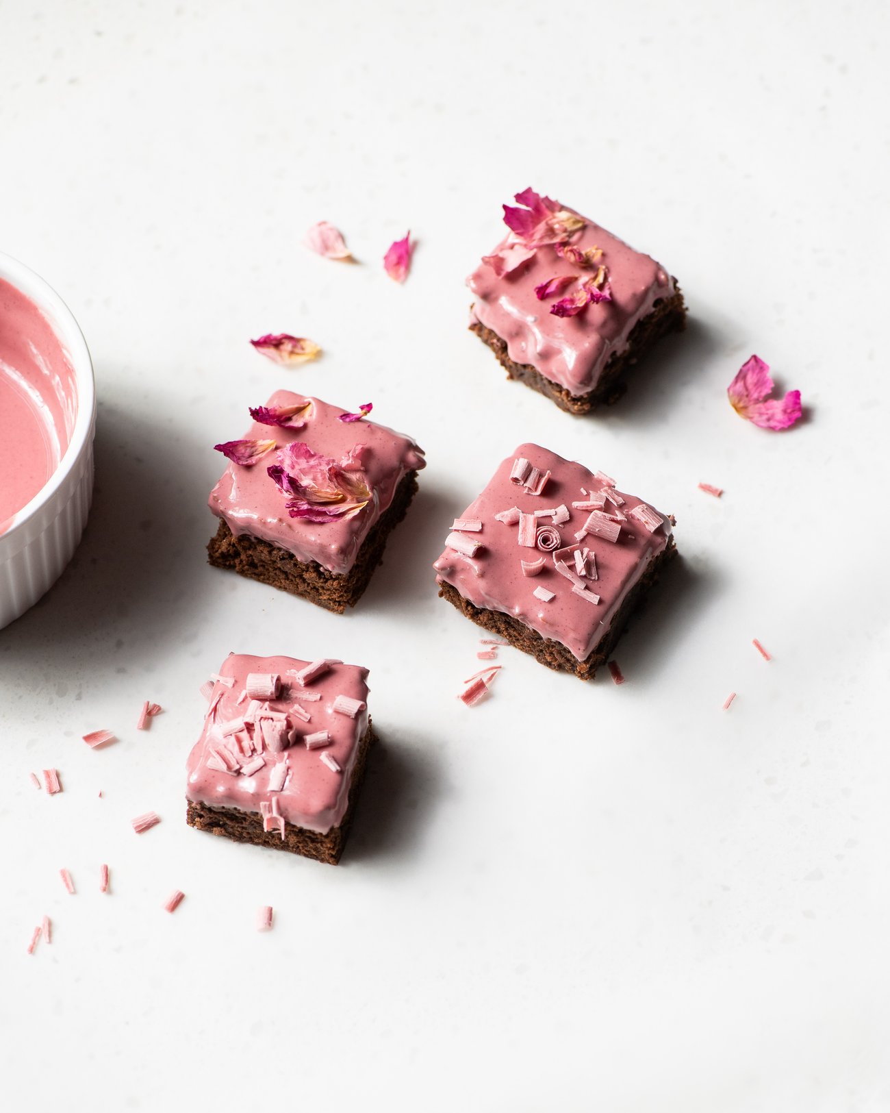 Top view of four brown cake squares on white counter topped with pink icing, pink chocolate shavings and pink flower petals