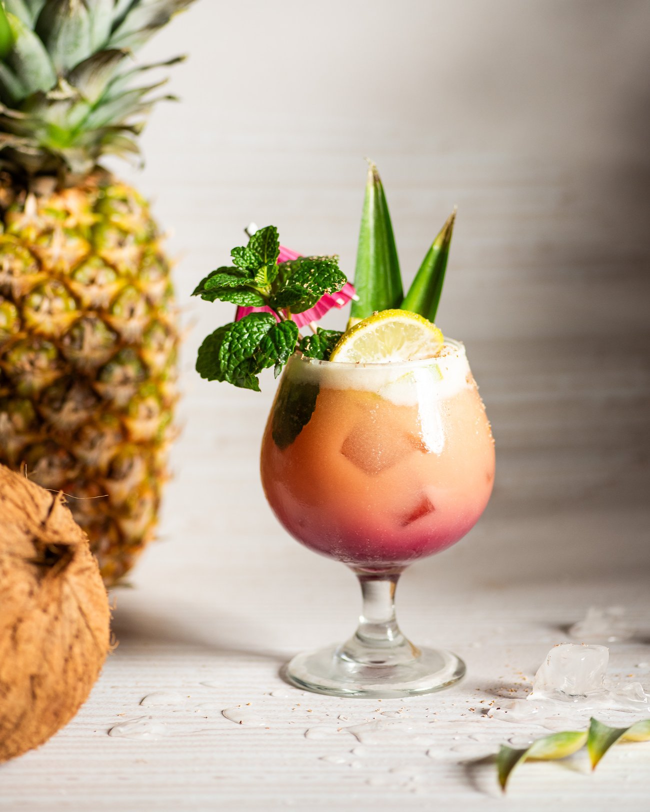 Colorful gradient pink to orange cocktail in large brandy glass with lemon and mint garnish next to a whole pineapple and coconut