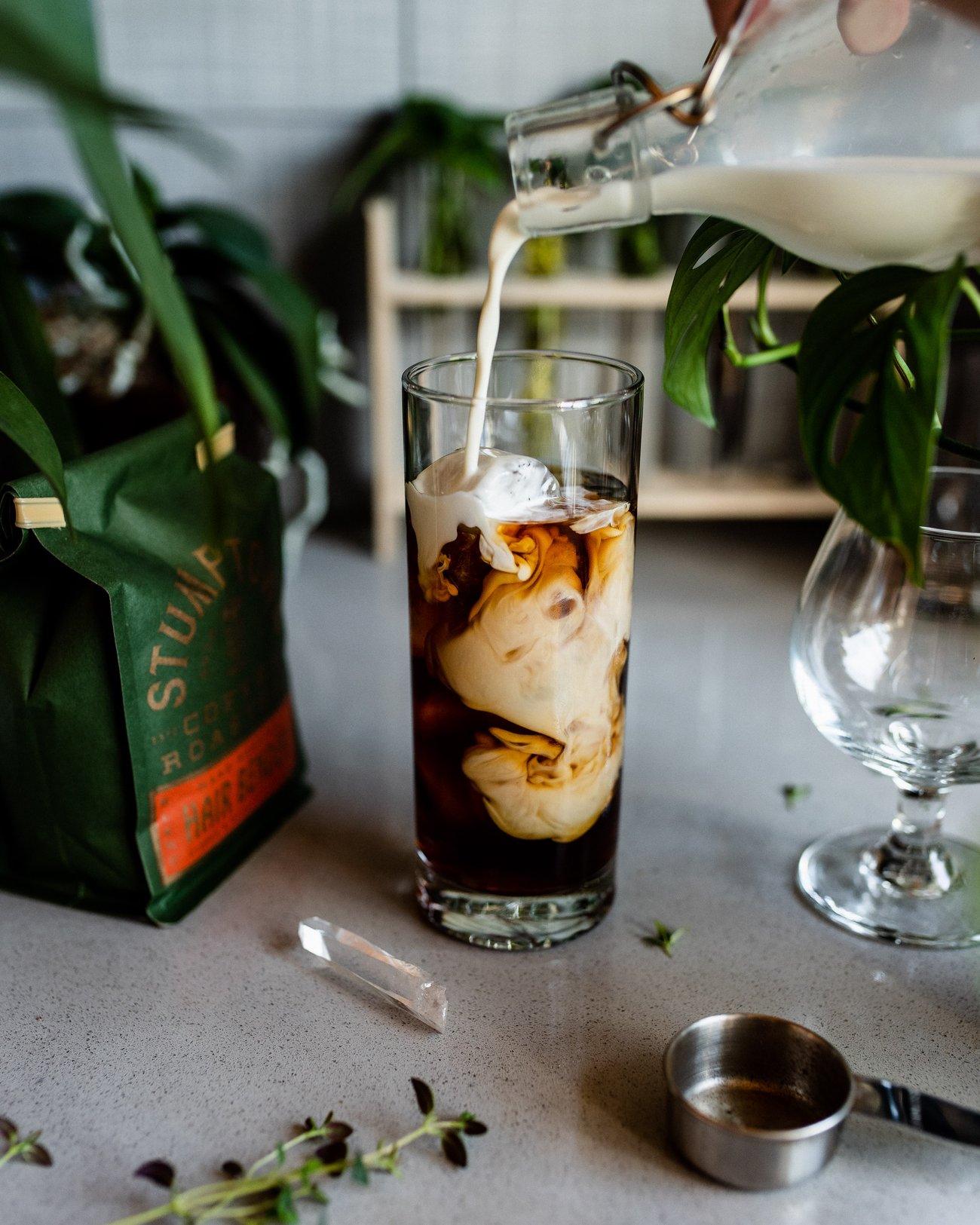 Milk swirling while being poured into tall glass of ice coffee on kitchen counter with bag of coffee beans, quartz crystal and plants
