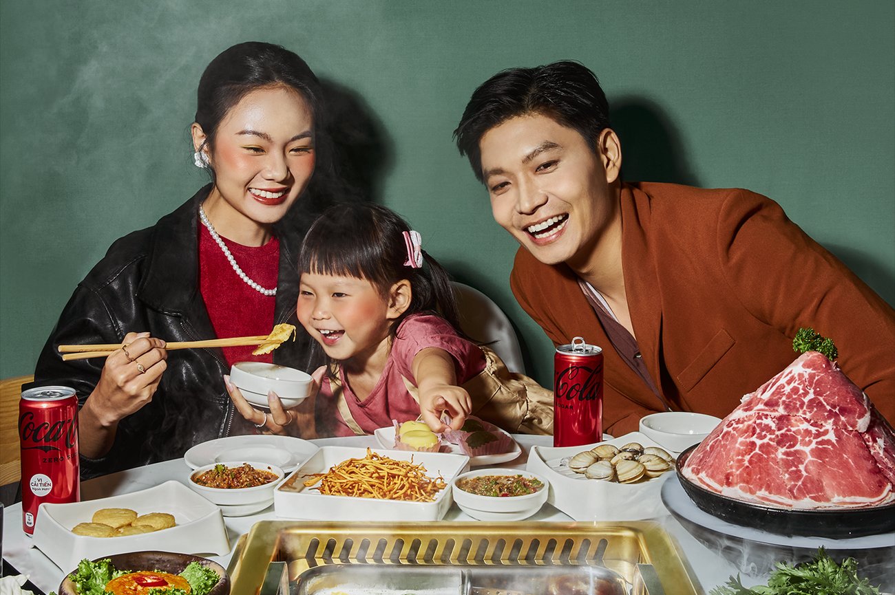 Smiling Asian mother, father and daughter eating steaming hot pot from table with large plates of meat, seafood, vegetables and noodles against a dark green wall