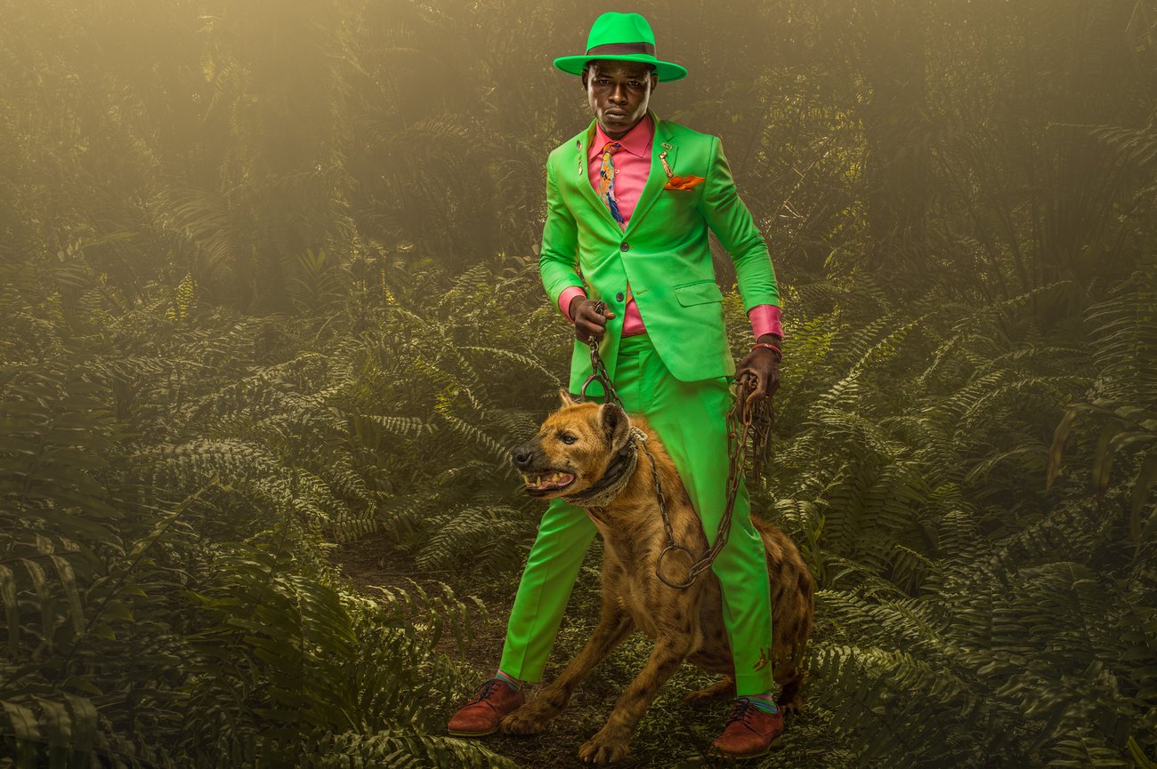 Black man in dense jungle in bright green suit and hat with pink shirt holding a snarling hyena with a metal chain