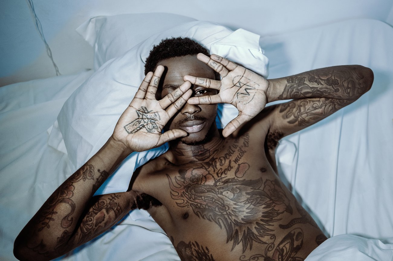 Tattooed Black man lying in white sheets with one eye looking at camera through fingers