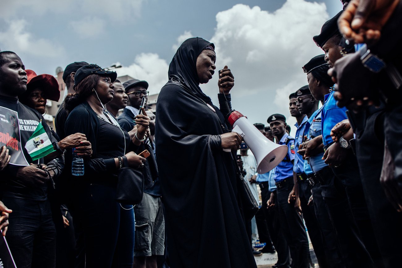 Black woman in headscarf holding megaphone addressing line of police in front of protesting crowd