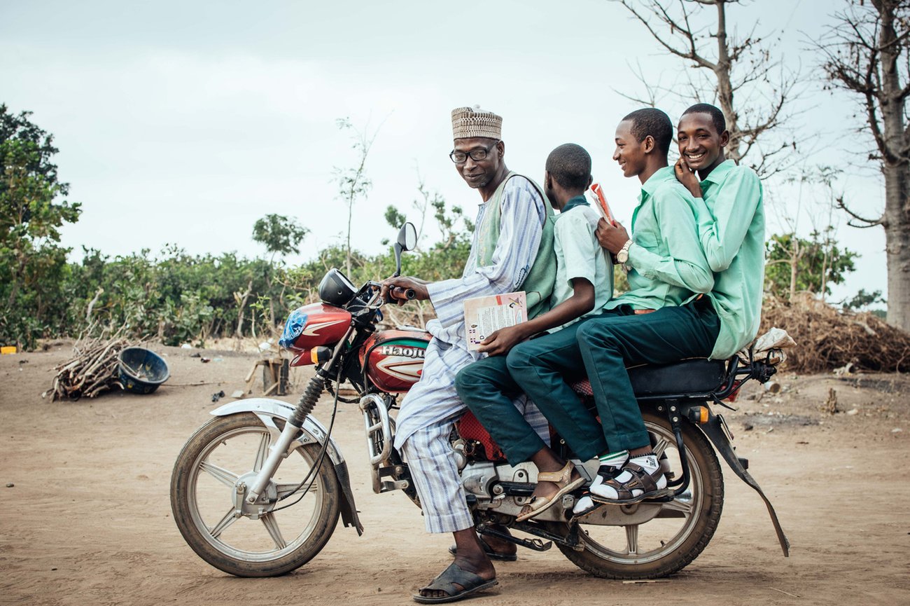 Black African man in Kufi hat and three boys dressed in matching green shirts on the back of a motorcycle