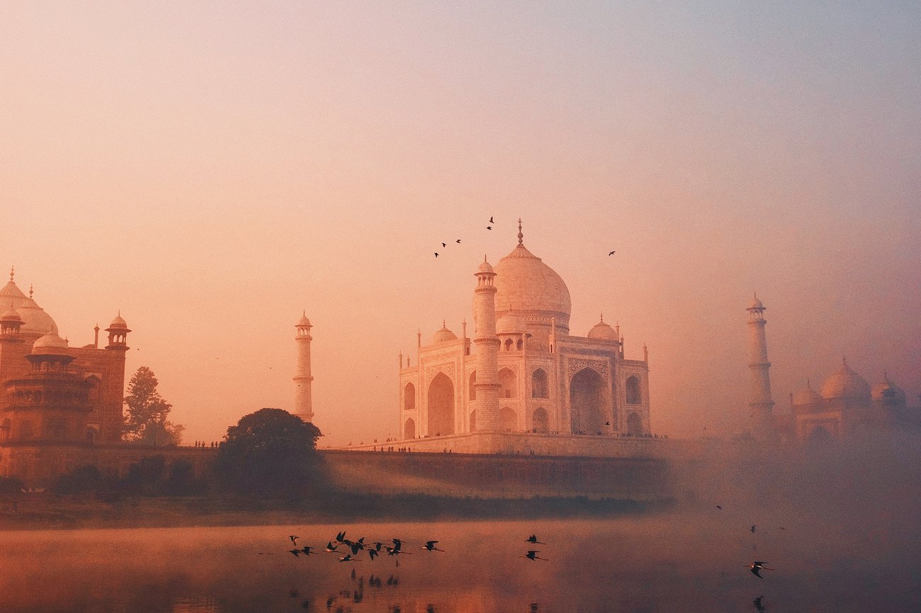 Flocks of birds flying in front of the Taj Mahal in India with peach and blue hues are seen from boat in the Yamuna river